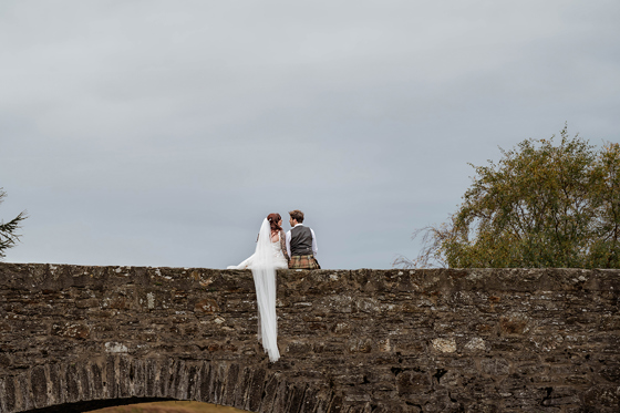 Bride and groom sitting on wall of stone bridge with veil hanging down